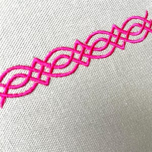 Op de afbeelding: Close-up van een lichtgrijze stof met een levendige roze geborduurde rand. De rand heeft een herhalend geometrisch ontwerp van verweven vormen, waardoor een decoratief element ontstaat. De borduursels voegen kleur en textuur toe aan de stof.