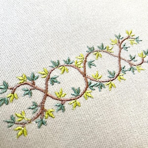Puede incluir: Diseño bordado de una vid con hojas verdes y amarillas sobre un fondo de tela blanca.