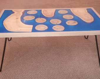 May include: A wooden coffee table with a blue epoxy resin inlay and a unique design featuring wood circles. The table has black hairpin legs.