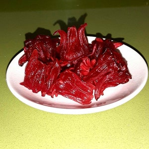 Może przedstawiać: Biały talerz z kupką suszonych czerwonych kwiatów hibiskusa. Kwiaty są ułożone luźno i mają intensywny czerwony kolor.