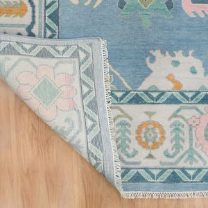 K&ouml;nnte beinhalten: Ein hellblauer Teppich mit einem wei&szlig;en und hellblauen Muster. Der Teppich hat einen Fransenrand und zeigt ein florales Motiv.
