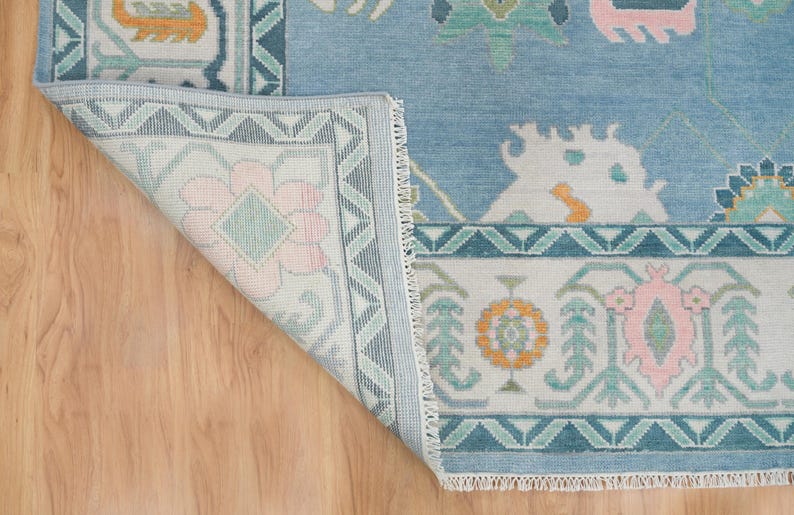 K&ouml;nnte beinhalten: Ein hellblauer Teppich mit einem wei&szlig;en und hellblauen Muster. Der Teppich hat einen Fransenrand und zeigt ein florales Motiv.