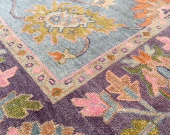 Könnte beinhalten: Nahaufnahme eines gemusterten Teppichs mit hellblauem Hintergrund und violetter Umrandung. Der Teppich zeigt ein florales Design mit rosa, orangefarbenen und grünen Akzenten.