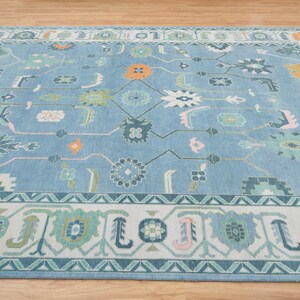 K&ouml;nnte beinhalten: Ein blauer Teppich mit einem wei&szlig;en, rosa und gr&uuml;nen geometrischen Muster. Der Teppich hat einen Rand mit einem sich wiederholenden Muster aus Dreiecken und Blumen.