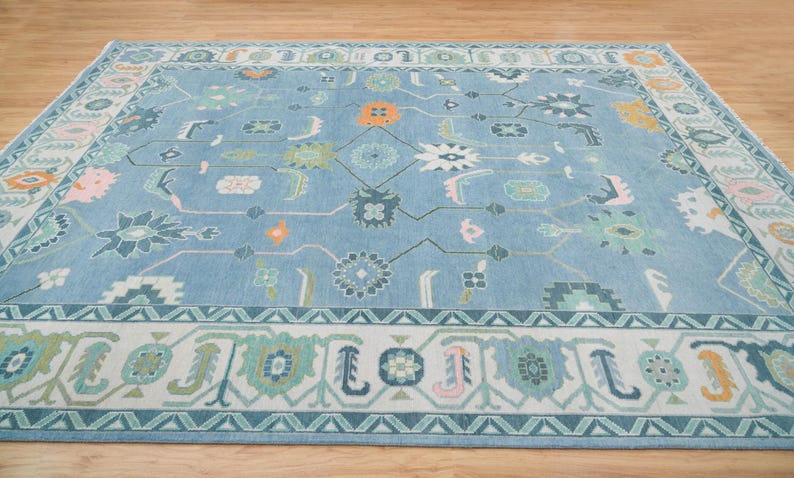 K&ouml;nnte beinhalten: Ein blauer Teppich mit einem wei&szlig;en, rosa und gr&uuml;nen geometrischen Muster. Der Teppich hat einen Rand mit einem sich wiederholenden Muster aus Dreiecken und Blumen.