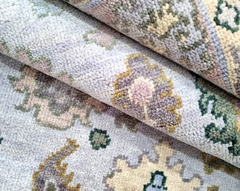 May include: Close-up of a rolled-up area rug with a floral pattern. The rug features a light blue background with detailed designs in shades of beige, green, and purple. The texture appears soft and plush, suggesting a comfortable floor covering.