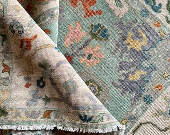 May include: Rolled-up area rug with a detailed floral pattern in teal, beige, orange, and pink. The rug has a fringed edge and a vintage, distressed appearance, showcasing intricate designs and textures.