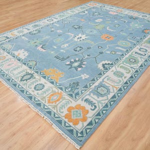 K&ouml;nnte beinhalten: Ein blauer Teppich mit einem wei&szlig;en und gr&uuml;nen Rand. Der Teppich hat ein Blumenmuster mit orangefarbenen, rosa und gr&uuml;nen Akzenten.