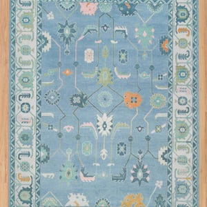 K&ouml;nnte beinhalten: Ein blauer Teppich mit einem geometrischen Muster in Rosa-, Gr&uuml;n-, Orange- und Wei&szlig;t&ouml;nen. Der Teppich hat einen Rand mit einem sich wiederholenden Muster.