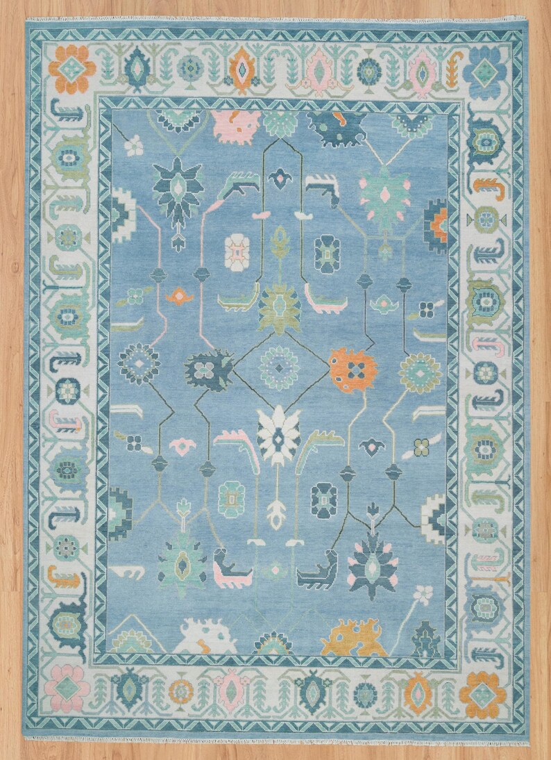 K&ouml;nnte beinhalten: Ein blauer Teppich mit einem geometrischen Muster in Rosa-, Gr&uuml;n-, Orange- und Wei&szlig;t&ouml;nen. Der Teppich hat einen Rand mit einem sich wiederholenden Muster.