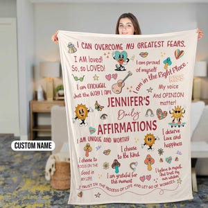 May include: A beige throw blanket with colorful illustrations and text. The text includes affirmations such as "I am loved!" and "I am enough." The blanket also features a personalized name, "Jennifer's Daily Affirmations".