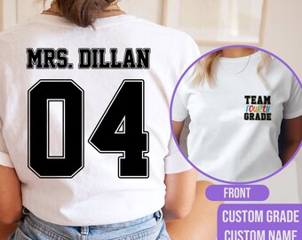 Op de afbeelding: Wit T-shirt met zwarte tekst "MRS. DILLAN 04" op de achterkant. De voorkant van het shirt heeft de tekst "TEAM FOURTH GRADE" in regenboogkleuren.