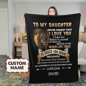 May include: Black fleece blanket with a lion illustration and text that reads "TO MY DAUGHTER, I LOVE YOU, LOVE, DAD". The blanket has a customizable area for a name. The blanket is held up by a person.