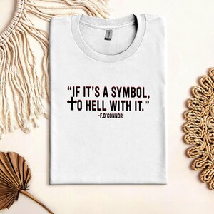 May include: White t-shirt with the quote "IF IT'S A SYMBOL, TO HELL WITH IT." in red and black text. The quote is attributed to F.O'Connor. The shirt is folded neatly on a light-colored surface, with decorative elements in the background.