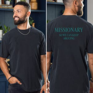 May include: Black short-sleeve t-shirt with the word "MISSIONARY" and the phrase "SO WE CAN KEEP ARGUING" printed on the back in teal. The shirt is a relaxed fit and has a crew neck.