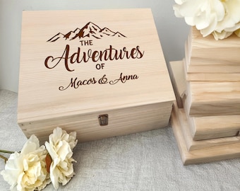 May include: A light-coloured wooden box featuring a mountain range design and the words "The Adventures of Macos & Anna" engraved on the lid. The box is secured with a small metal clasp. Flowers are in the foreground.