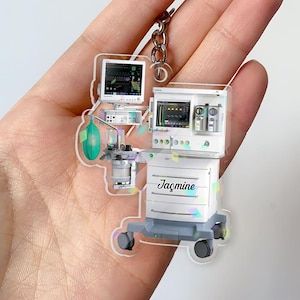 Puede incluir: Un llavero acrílico transparente con una máquina médica en miniatura con un monitor, medidores y una bolsa de respiración. La máquina es blanca con detalles grises y tiene el nombre "Jasmine" impreso. El llavero tiene una cadena plateada.