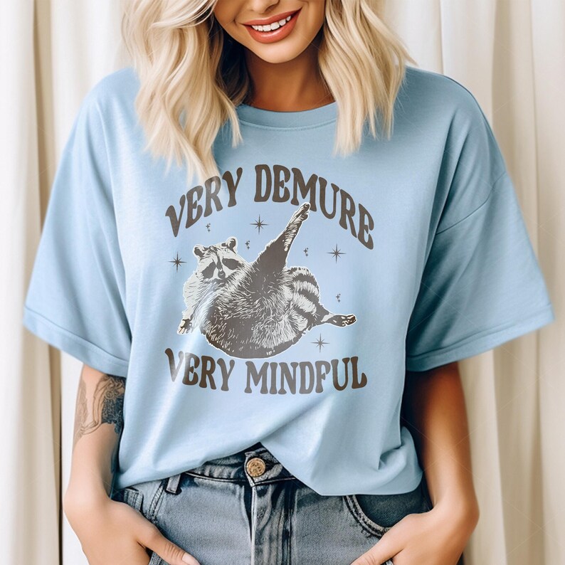 May include: A light blue t-shirt featuring a graphic of a raccoon with the text "Very Demure Very Mindful".