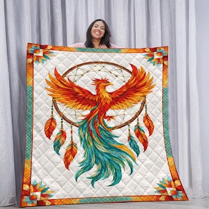 May include: A white quilted blanket featuring a vibrant phoenix design within a dreamcatcher. The phoenix is depicted with orange, red, and teal feathers. The blanket has a decorative border with geometric patterns and feather accents.