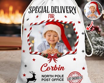 May include: White Christmas gift bag with a drawstring closure. The bag features a photo of a child wearing a Santa hat, the text "SPECIAL DELIVERY FOR Corbin", and a reindeer graphic. Additional text includes "NORTH POLE POST OFFICE" and "LIST CHECKED 2X".
