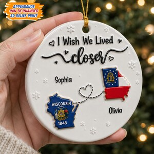 Op de afbeelding: Een ronde witte keramische hanger met de tekst I Wish We Lived closer, 3D-reliëfkaarten van Wisconsin en Georgia verbonden door een stippellijn in de vorm van een hart. De namen Sophia en Olivia staan bij de betreffende staten vermeld.