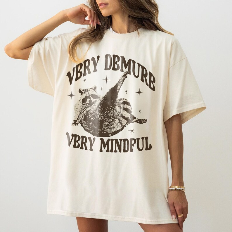 May include: An oversized cream-coloured t-shirt with a brown graphic of a raccoon with the text "Very Demure Very Mindful".