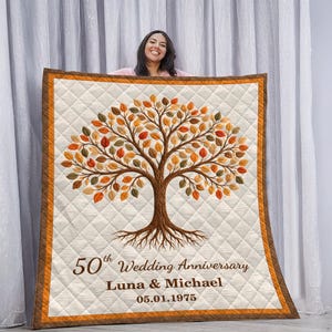Könnte beinhalten: Eine cremefarbene Steppdecke mit einem Baumdesign und Herbstblättern. Sie hat einen braunen Rand und den Text "50th Wedding Anniversary Luna & Michael 05.01.1975". Der Hintergrund ist ein weißer Vorhang.