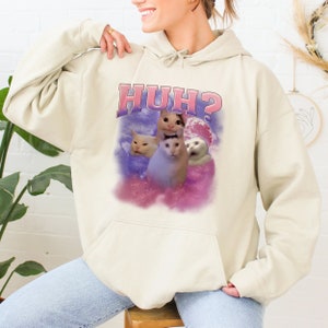 K&ouml;nnte beinhalten: Ein cremefarbener Hoodie mit einem pink- und lilafarbenen Aufdruck von drei Katzen und dem Text "HUH?" auf der Vorderseite.