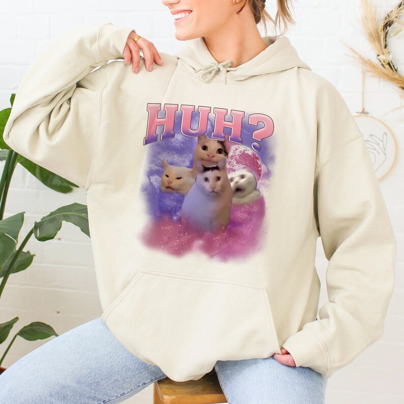K&ouml;nnte beinhalten: Ein cremefarbener Hoodie mit einem pink- und lilafarbenen Aufdruck von drei Katzen und dem Text "HUH?" auf der Vorderseite.