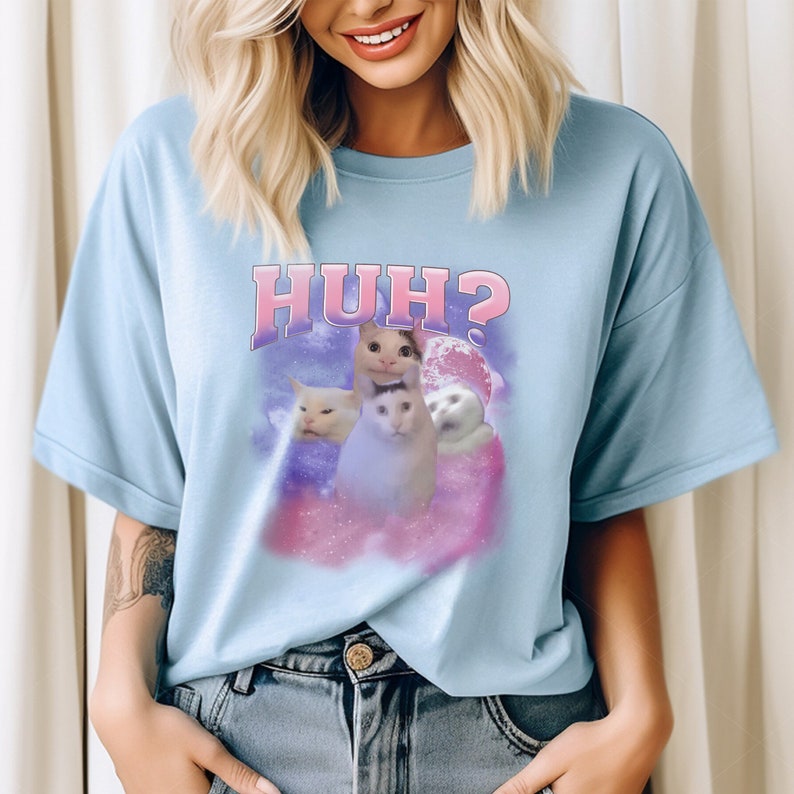 K&ouml;nnte beinhalten: Ein hellblaues T-Shirt mit einem rosa und wei&szlig;en Aufdruck von Katzen mit dem Text "HUH?" darauf.