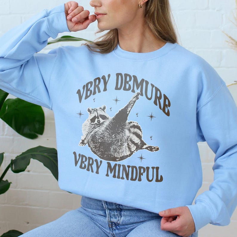 May include: A light blue sweatshirt featuring a raccoon graphic and the text "Very Demure Very Mindful".