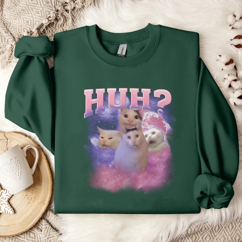 K&ouml;nnte beinhalten: Ein dunkelgr&uuml;ner Pullover mit einem rosa und wei&szlig;en Aufdruck von Katzen mit dem Text "HUH?" auf der Vorderseite.