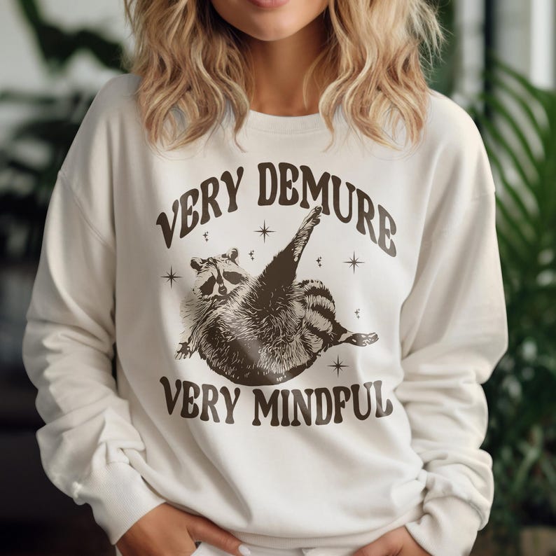 May include: A white sweatshirt with a brown raccoon graphic and the text "Very Demure Very Mindful".