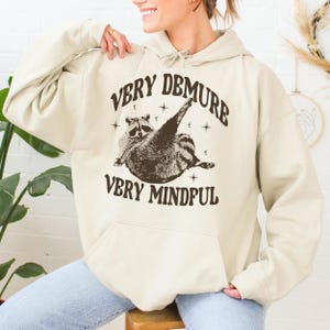 May include: A cream-coloured hoodie with a brown raccoon graphic and the text "Very Demure Very Mindful".