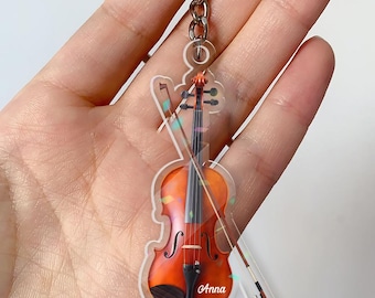 Puede incluir: Un llavero acrílico transparente con forma de violín con arco. El violín es naranja y marrón con cuerdas negras. El nombre "Anna" está impreso en la parte inferior. Una cadena y un anillo plateados están unidos en la parte superior.