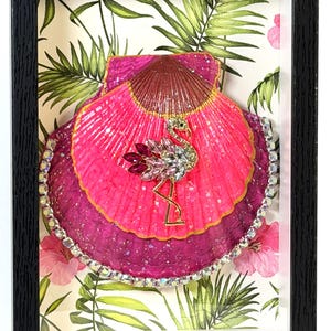 May include: A shadow box frame containing a layered scallop shell art piece. The shell is painted in vibrant shades of hot pink and magenta, adorned with a gold-toned flamingo brooch featuring pink and clear crystals, and edged with clear rhinestones.