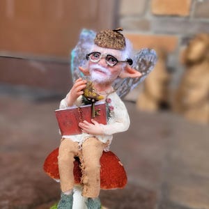 Peut inclure: Figurine de fée fantaisiste avec une barbe blanche, des lunettes et un chapeau marron, tenant un livre rouge et un petit oiseau. La fée est assise sur un champignon rouge et blanc sur une base en bois avec de la mousse verte.