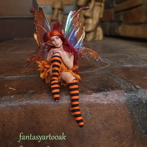 May include: A fairy figurine with long red hair, pointed ears, and iridescent copper wings. The figure wears a skirt of orange petals and black and orange striped stockings. The text "fantasyartooak Sep -2026" is printed in green at the bottom left.
