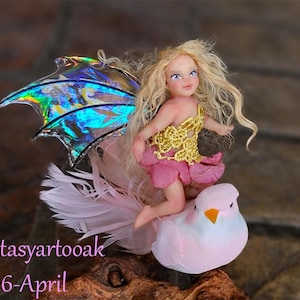 May include: A miniature fairy doll with blonde hair, a gold bodice, and a pink skirt, perched on a pink bird. The fairy has iridescent wings and is set against a blurred background. The text "fantasyartooak 2026-April" is visible.
