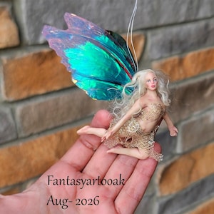 May include: A small fairy figurine with long blonde hair and a gold mesh bodice. The figure features large, iridescent wings that shift between blue and green tones. A thin white string is attached to the top of the wings for hanging purposes.