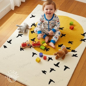 Könnte beinhalten: Cremefarbener Kinderteppich mit gelbem Sonnendesign und schwarzen Vogelsilhouetten. Ein lächelndes Baby sitzt auf dem Teppich, umgeben von Spielzeug, darunter eine Holzeisenbahn und bunte Blöcke. Der Teppich hat den Text "Tuftoria" in der Ecke.