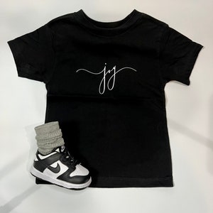 Könnte beinhalten: Schwarzes T-Shirt mit kurzen Ärmeln und dem Wort "joy" in weißer Kursivschrift.