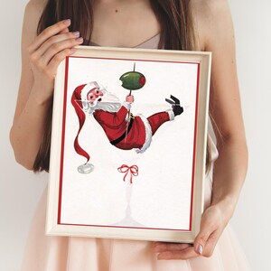 Op de afbeelding: Een wit ingelijste print van een cartoon kerstman die aan een martini-glas hangt met een olijf. De kerstman draagt een rood pak met witte versieringen en een witte baard. Het martini-glas is gevuld met een heldere vloeistof.