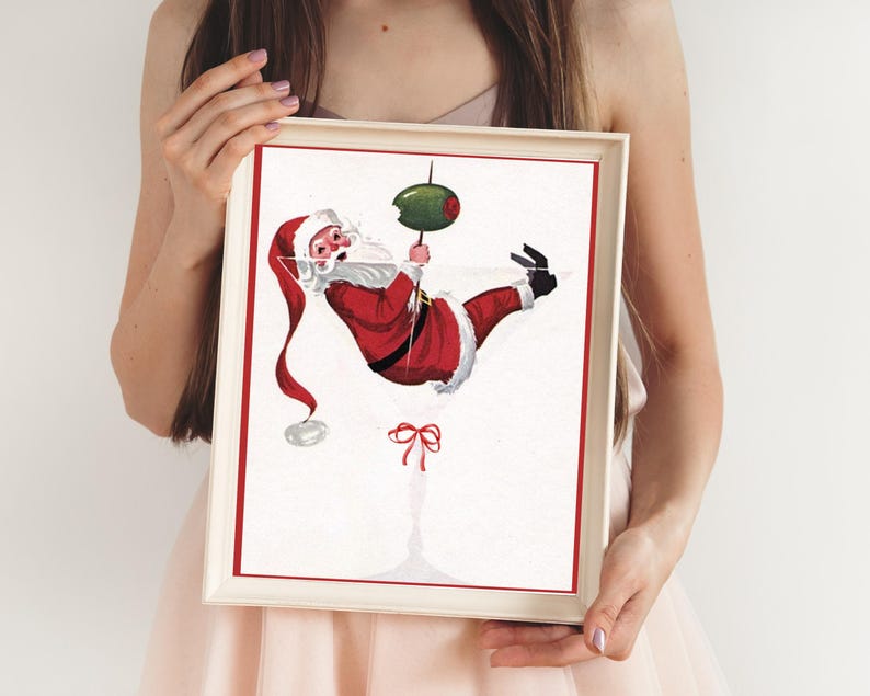 Op de afbeelding: Een wit ingelijste print van een cartoon kerstman die aan een martini-glas hangt met een olijf. De kerstman draagt een rood pak met witte versieringen en een witte baard. Het martini-glas is gevuld met een heldere vloeistof.