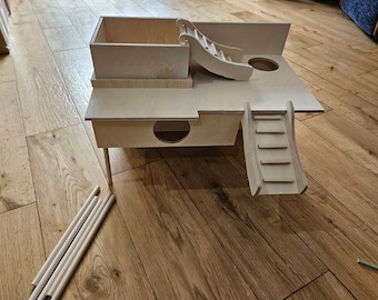 May include: A wooden hamster house with a ramp, a slide, and a platform. The house is made of natural wood and has a hole for the hamster to enter. The house is on a wooden floor.
