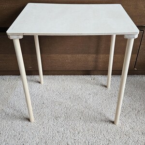 May include: A small, light-colored wooden table with a rectangular top and four rounded legs. The table is approximately 24 inches long, 18 inches wide, and 18 inches tall. The table is set on a light-colored carpet.