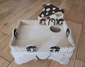 May include: A wooden playhouse for small animals, featuring a mushroom-shaped roof with black and white spots, a small door, and a circular opening. The playhouse is surrounded by a wooden fence with small wooden dowels.