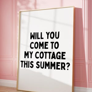 May include: A framed art print with the text "WILL YOU COME TO MY COTTAGE THIS SUMMER?" in black on a white background. The frame is gold, and the print leans against a pink wall with a wooden floor.