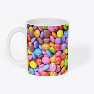 May include: White ceramic mug featuring a colourful design of assorted chocolate sweets. The sweets are in various shades of brown, red, orange, yellow, green, blue, and purple.
