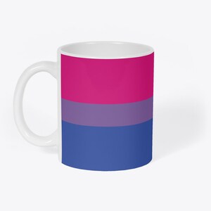 Może przedstawiać: Biały kubek ceramiczny z wzorem przedstawiającym flagę dumy biseksualnej. Flaga składa się z trzech poziomych pasów: różowego, fioletowego i niebieskiego.